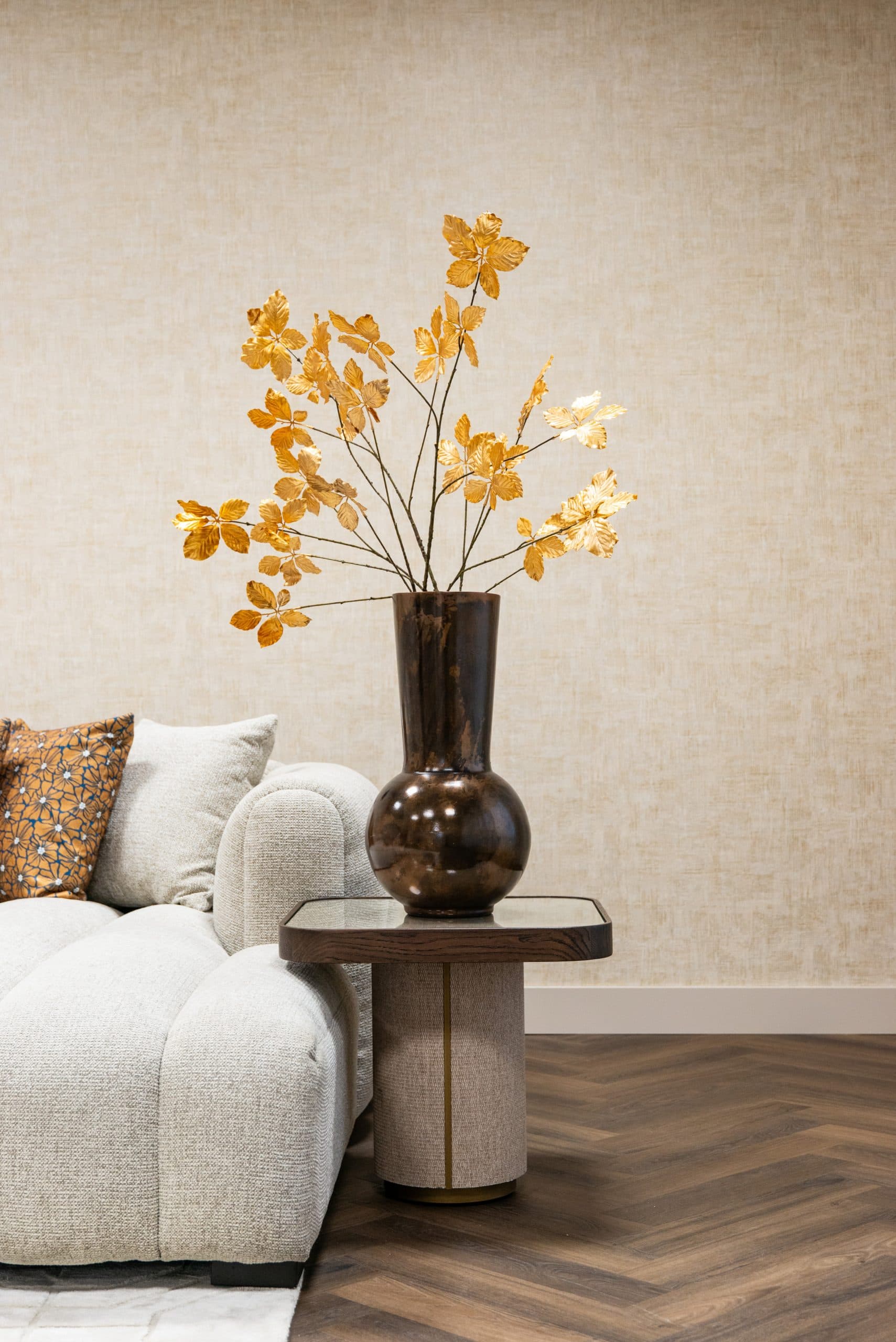 Hoge bronskleurige vaas met gouden takken naast een lichte bank met beige en okerkleurige kussens tegen een zandkleurige wand.