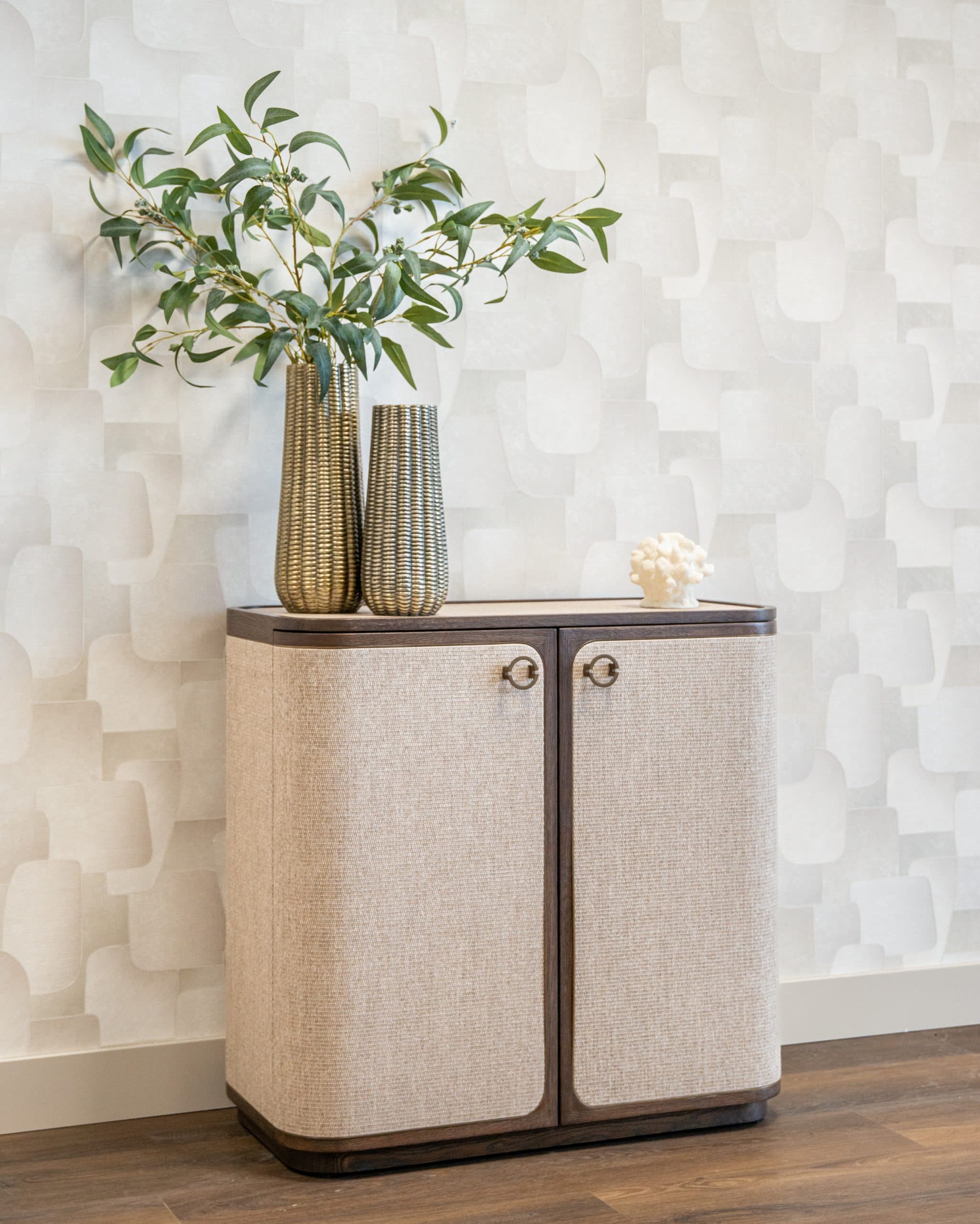 Beige dressoir met linnen deuren en ronde bronskleurige grepen, met twee goudkleurige geribbelde vazen en een wit koraalobject op het blad.