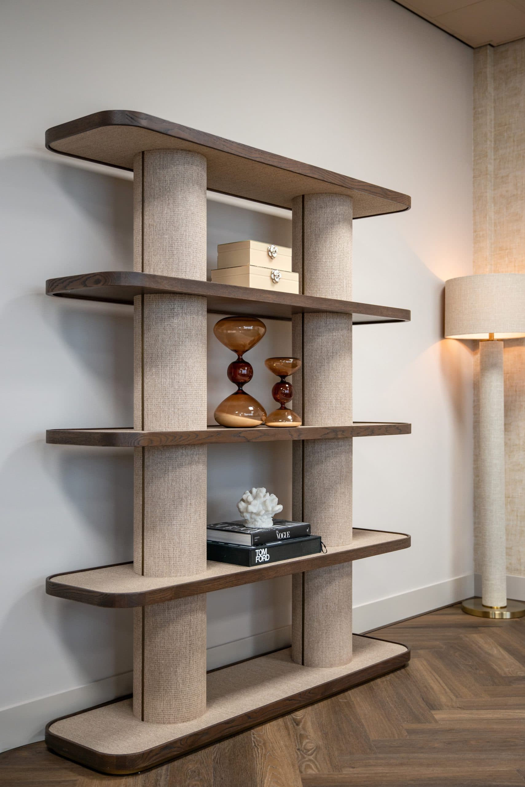Open kast met eiken planken en linnen pilaren, met twee glazen zandlopers, een witte koraaldecoratie en een opbergdoos, naast een brandende vloerlamp