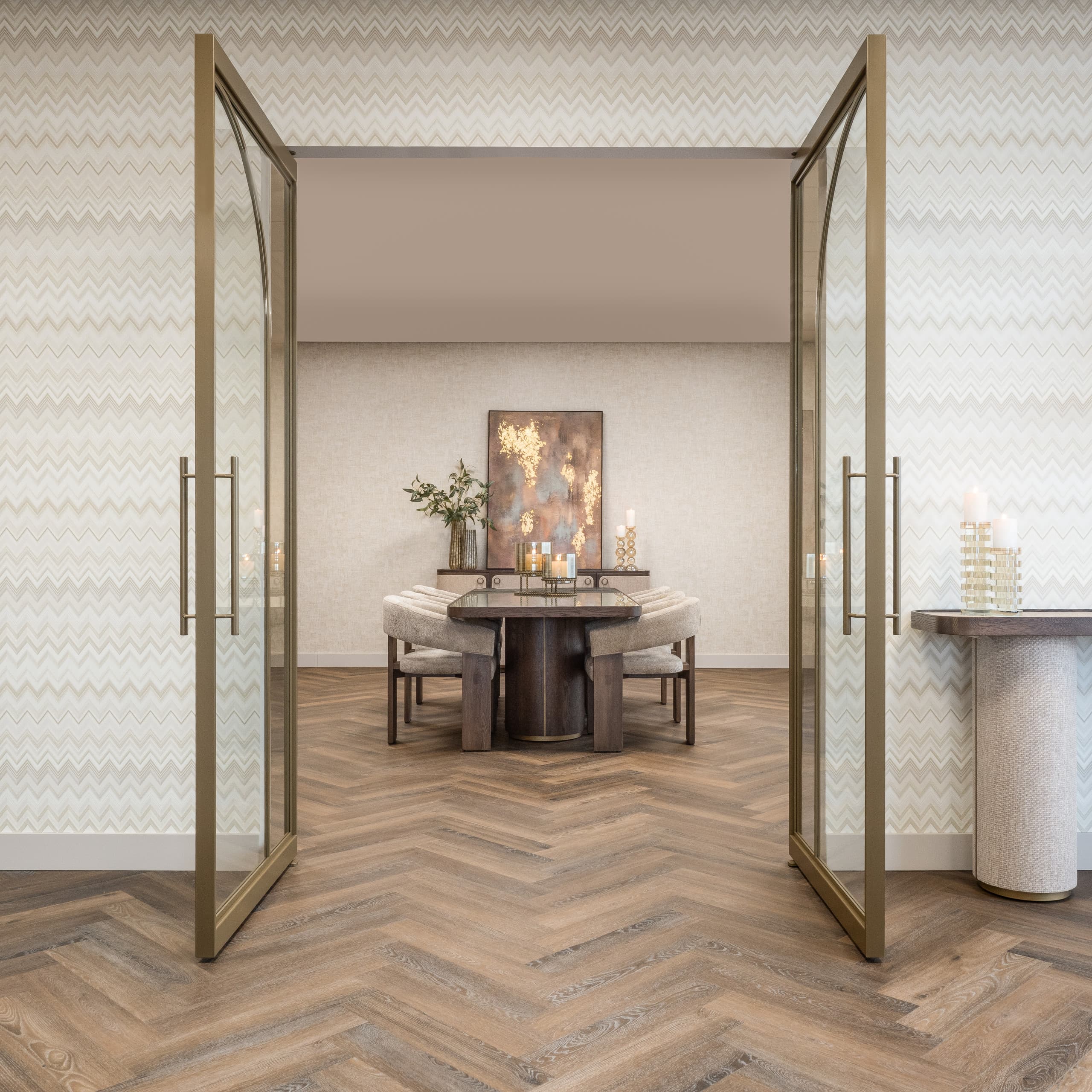 Blik door openstaande glazen deuren met messingkleurige lijsten op een eethoek met een ronde donkere tafel, lichte gestoffeerde stoelen en een schilderij aan de wand, op een visgraatvloer