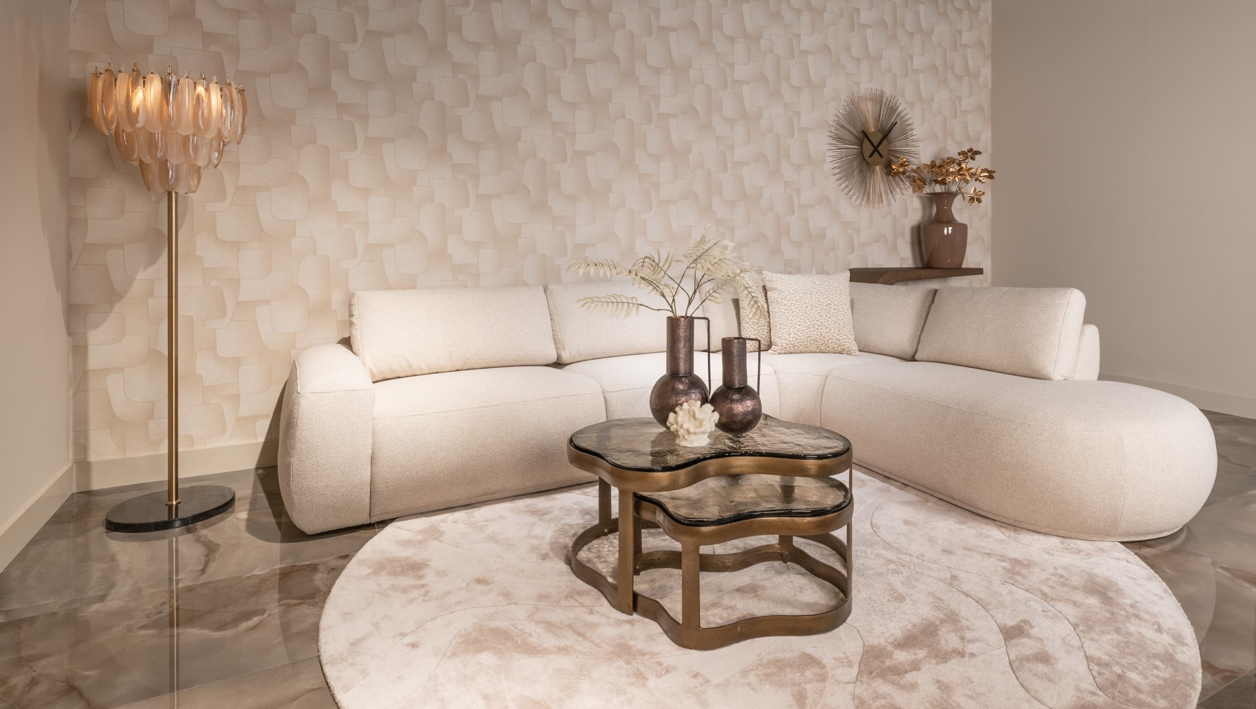 Woonkamer met een crèmekleurige gebogen bouclé hoekbank, twee bronskleurige salontafels met marmeren blad, een schelpvormige vloerlamp en een rond vloerkleed