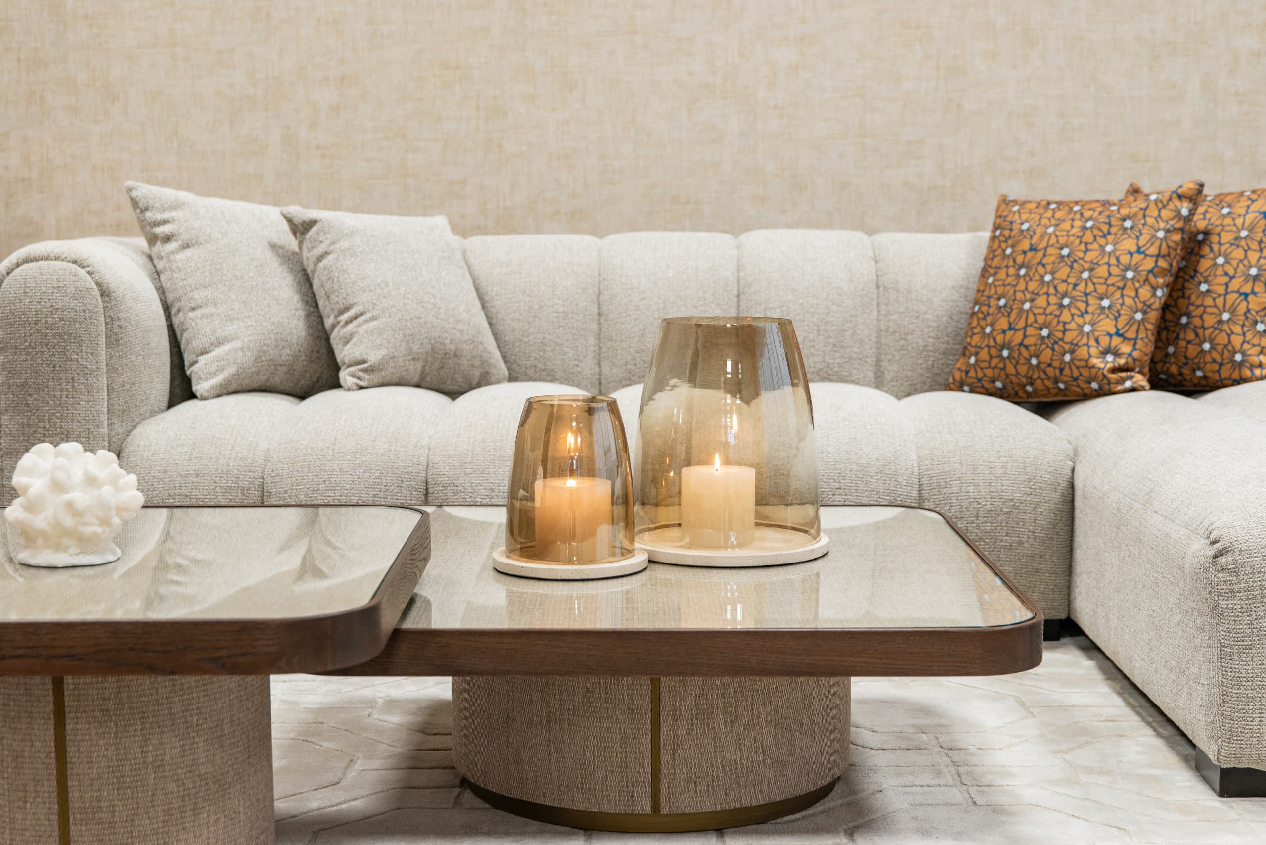 Twee brandende kaarsen in amberkleurige glazen windlichten op een houten salontafel, met een lichte hoekbank en kussens op de achtergrond