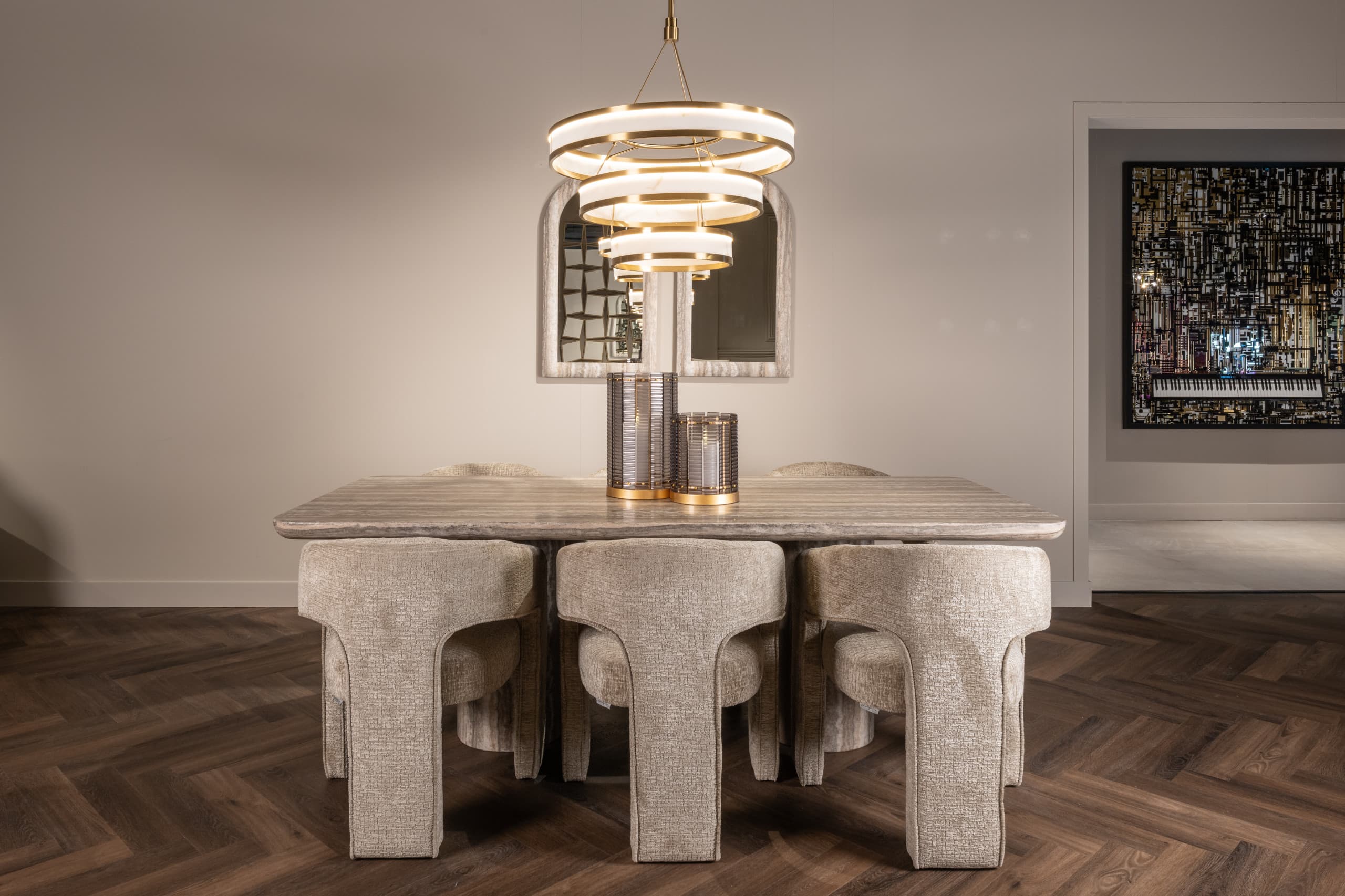 Eettafel met stoffen stoelen onder een verlichte hanglamp met meerdere ringen, met een boogspiegel aan de wand en windlichten op tafel