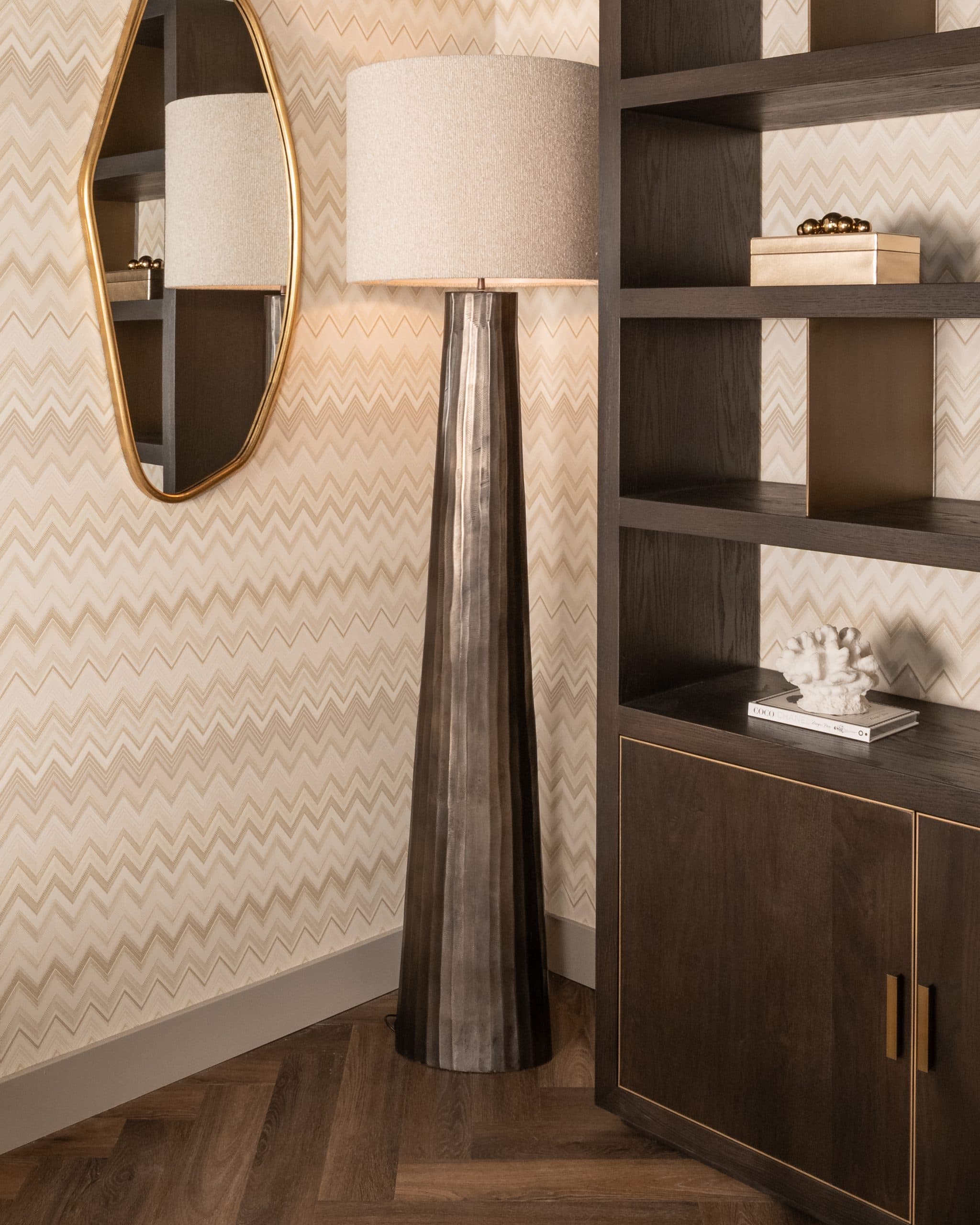 Hoge wand met zigzagbehang, een ovale gouden spiegel, een bronzen vloerlamp met beige kap en een donkere open kast met decoratie