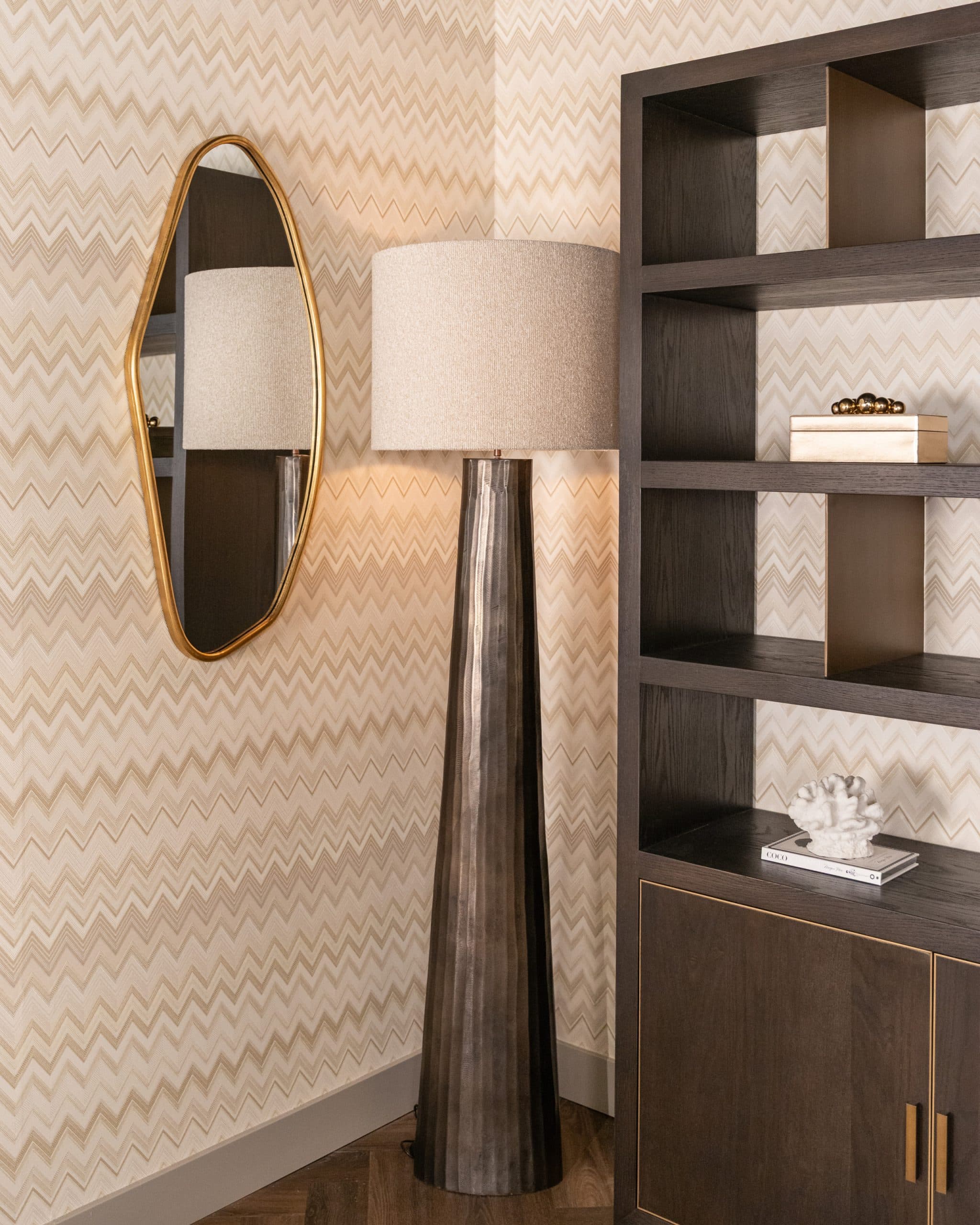Brandende lamp met linnen kap tegen een lichte wand met zigzagpatroon, naast een gouden spiegel en een donkere open kast met accessoires