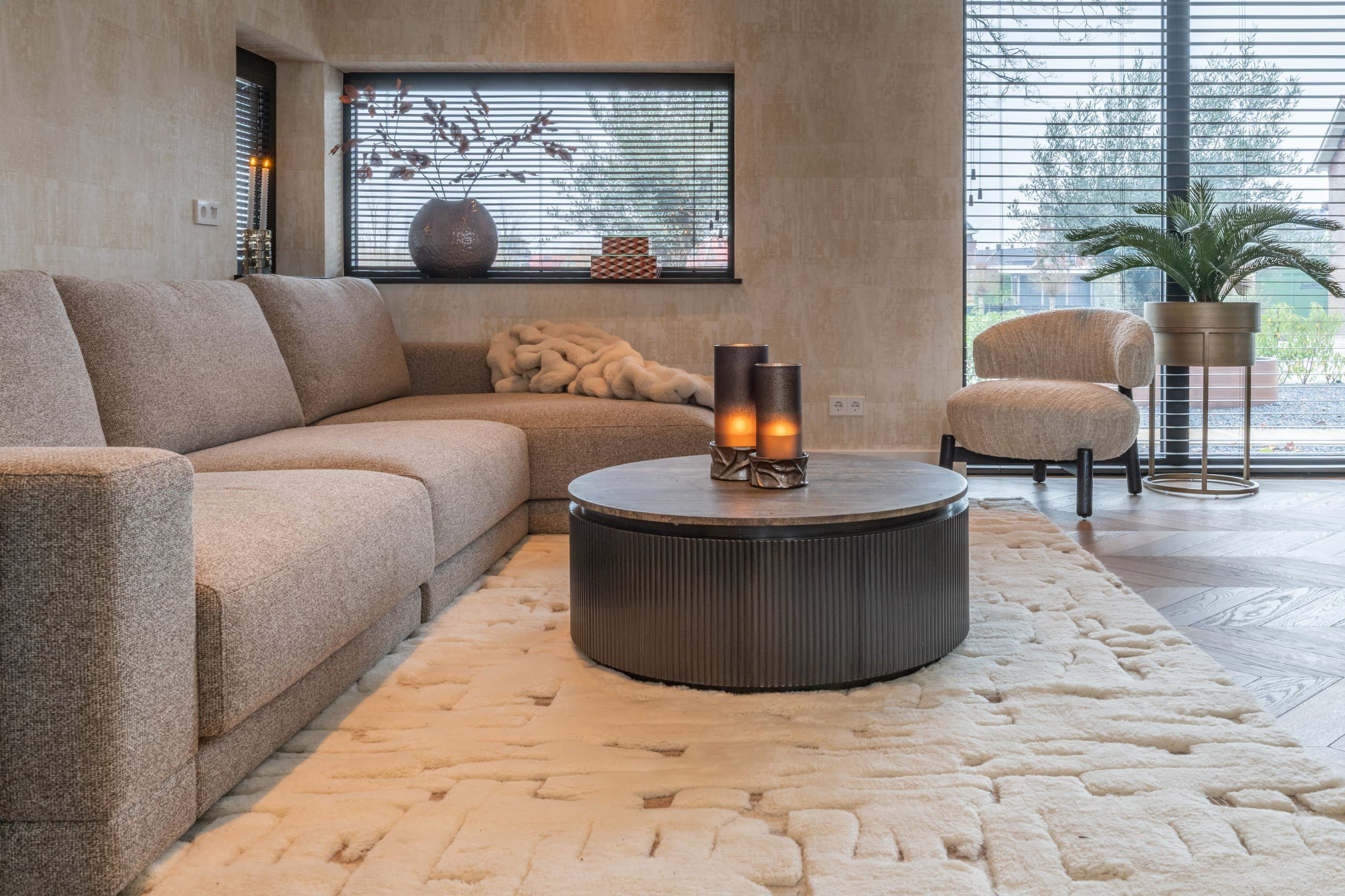 Beige hoekbank in een woonkamer met een ronde donkere salontafel, een bouclé fauteuil en een wollig vloerkleed