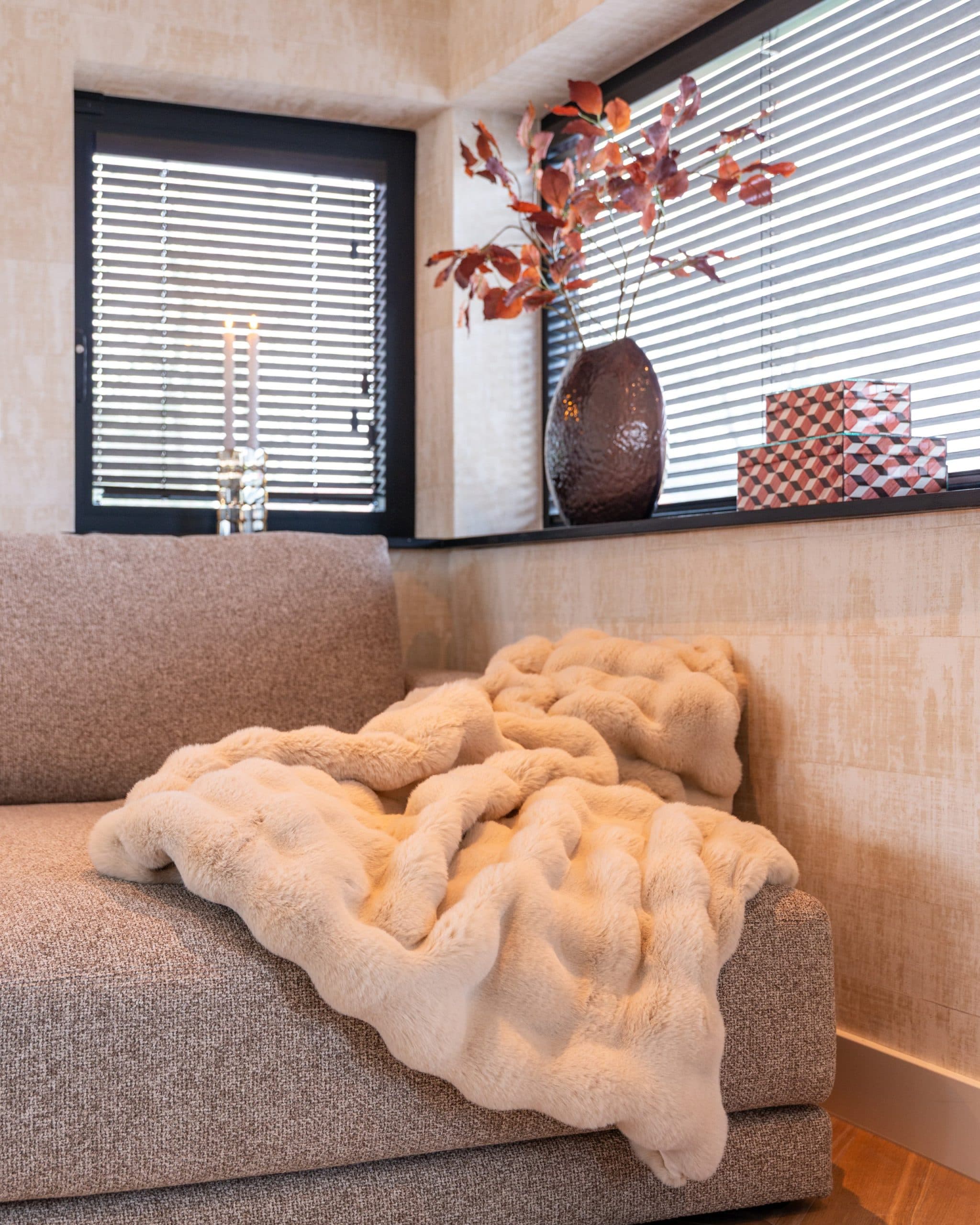 Hoek van een lichte bank met een zachte beige plaid, voor een vensterbank met een donkere vaas en decoratieve dozen