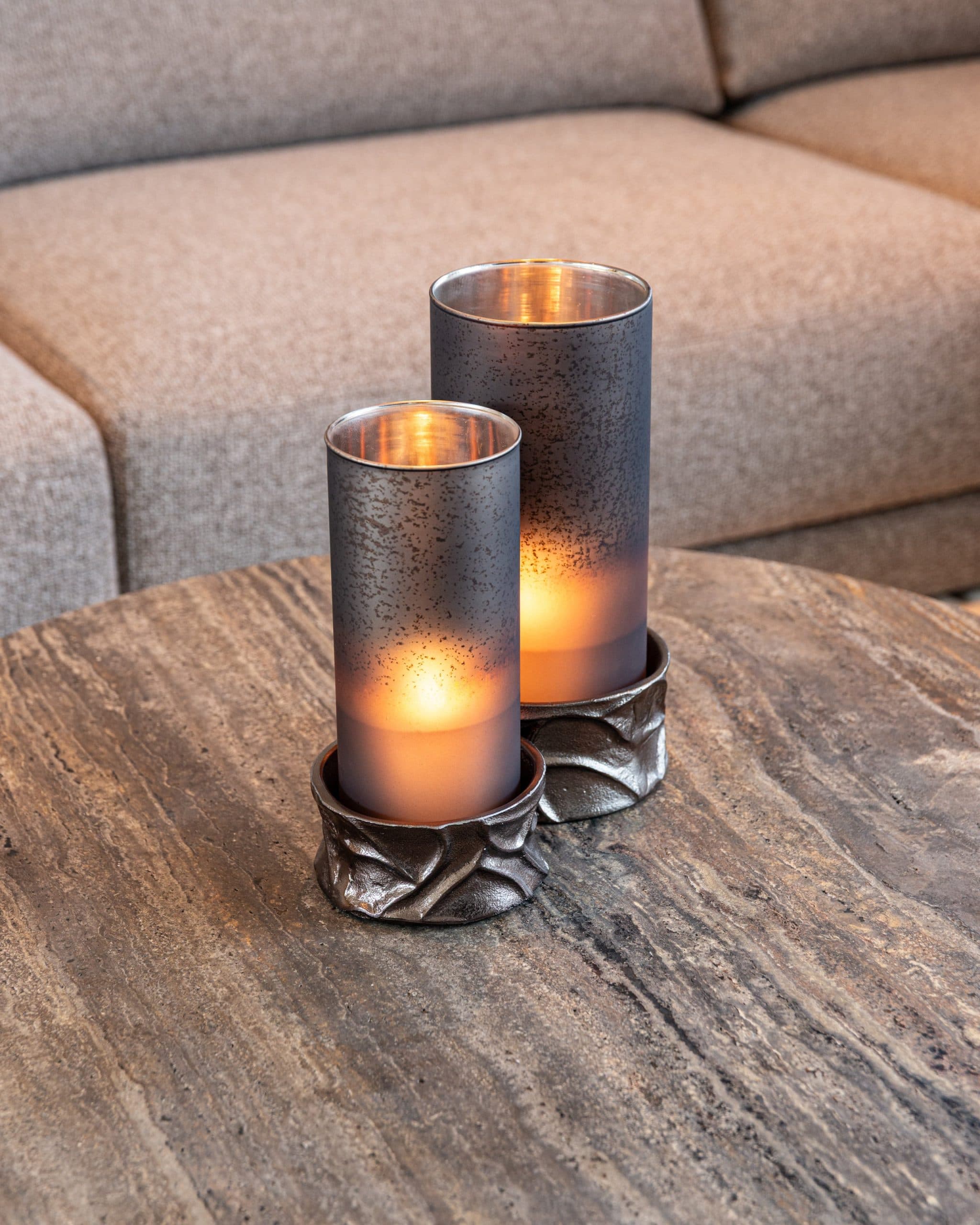 Twee brandende windlichten met gevlekt glas op ruwe metalen voetjes op een ronde travertijnen salontafel
