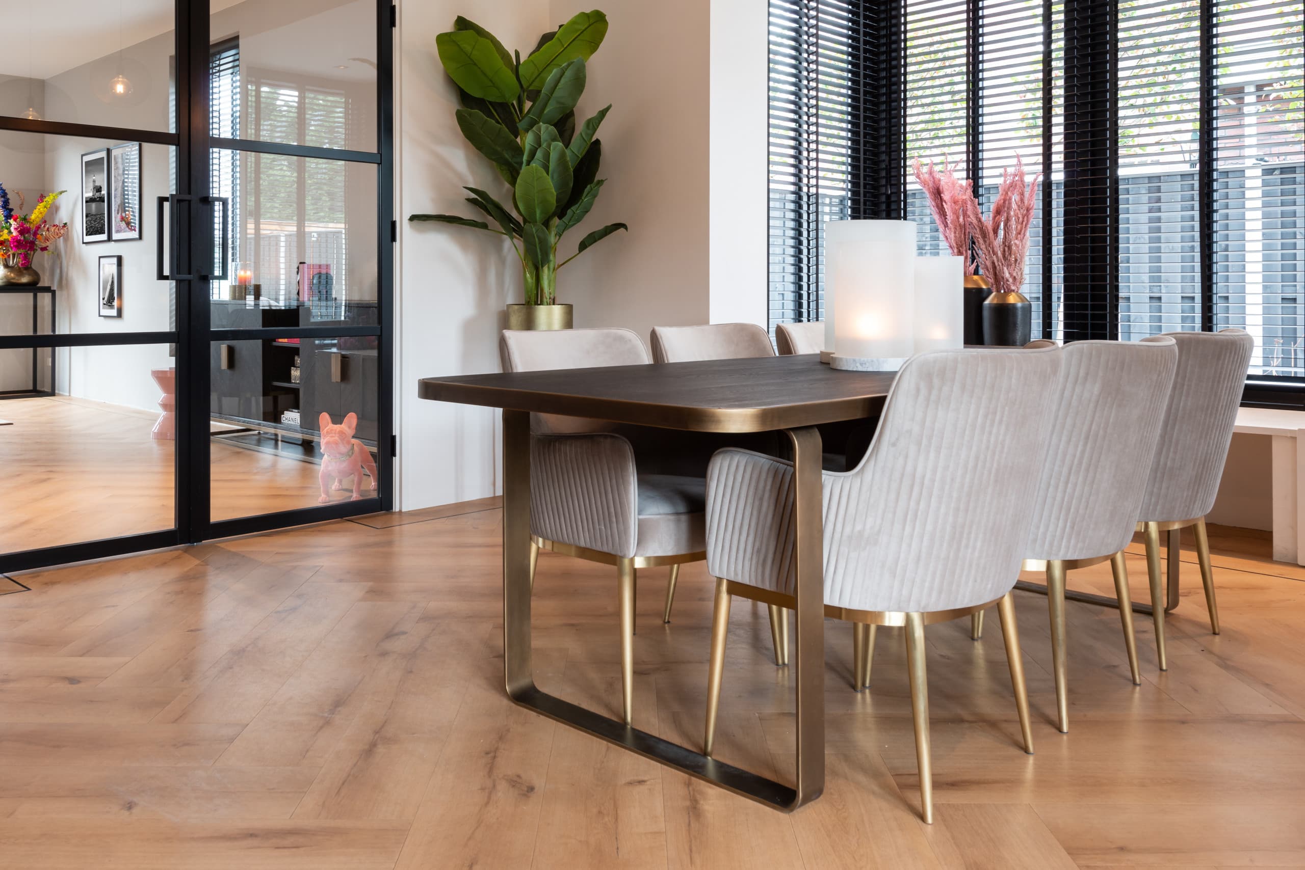 Eethoek met een donkere houten eettafel op een bronskleurig onderstel en zes gestoffeerde armstoelen in khaki velvet met gouden poten, naast een grote plant en een windlicht
