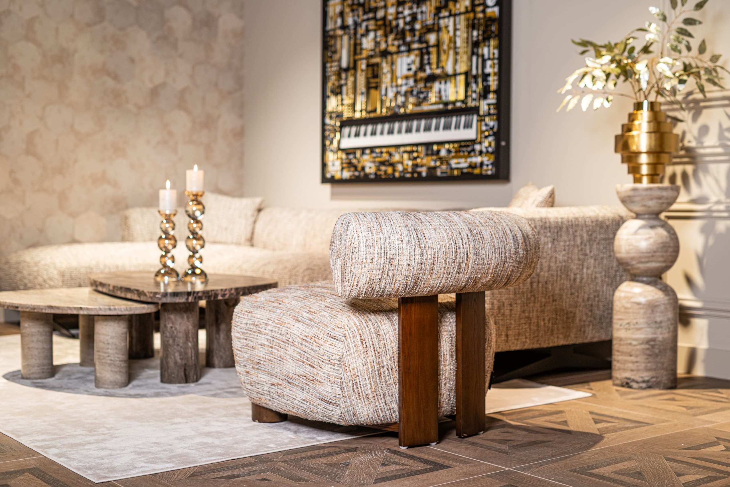 Woonkamer met een travertijn pilaar waarop een gouden vaas met olijftakken staat, naast een bouclé fauteuil en ronde salontafels.