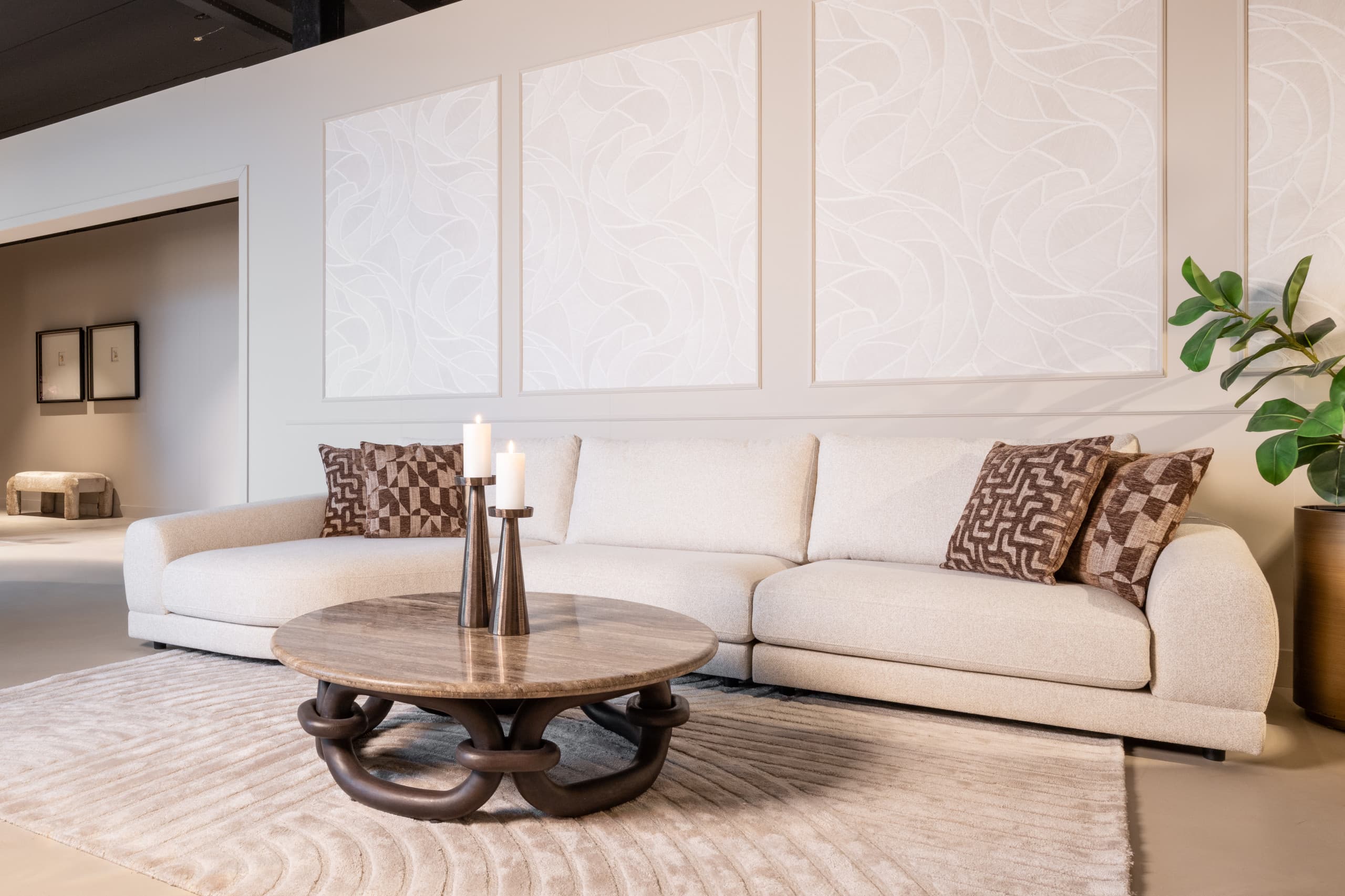 Ruime woonkamer met een crèmekleurige loungebank, bruin gedessineerde sierkussens, een ronde travertine salontafel met twee donkere kandelaars en witte wandpanelen erachter