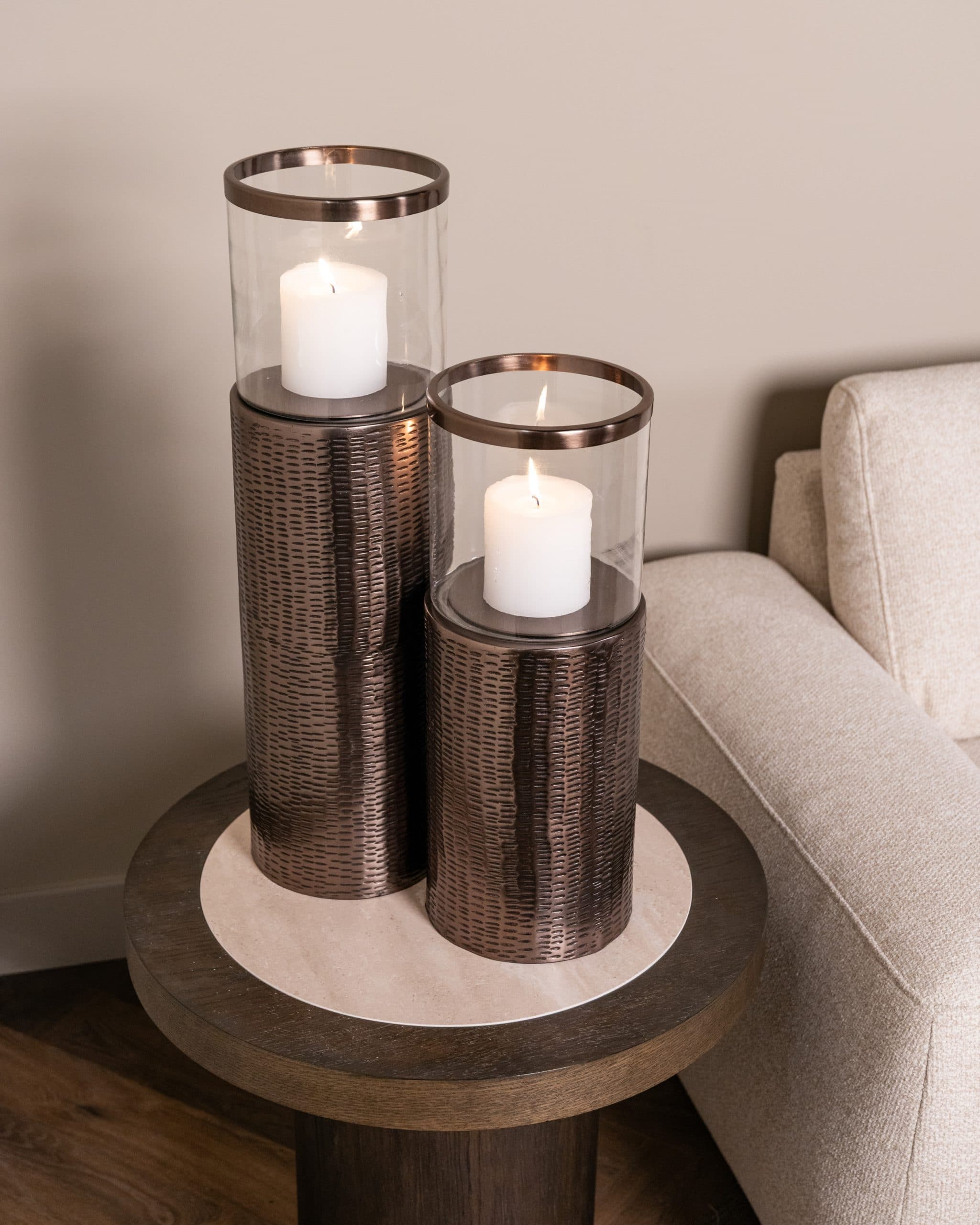 Twee bronskleurige windlichten met brandende stompkaarsen op een ronde bijzettafel naast een lichte fauteuil