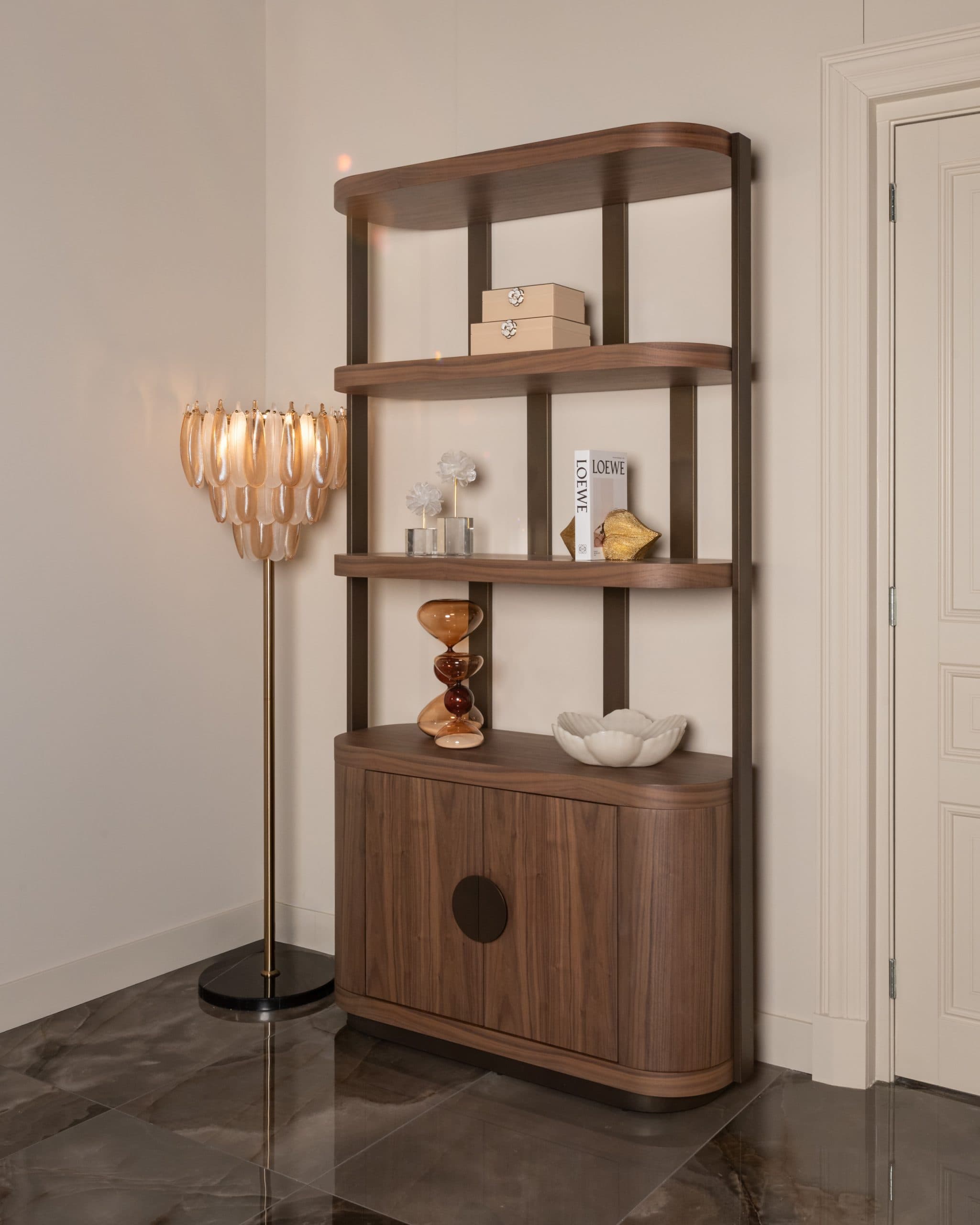 Open wandkast in walnoot tegen een lichte wand, met boeken, een goudkleurig object, glazen zandlopers en een witte schaal op de planken, naast een schemerlamp