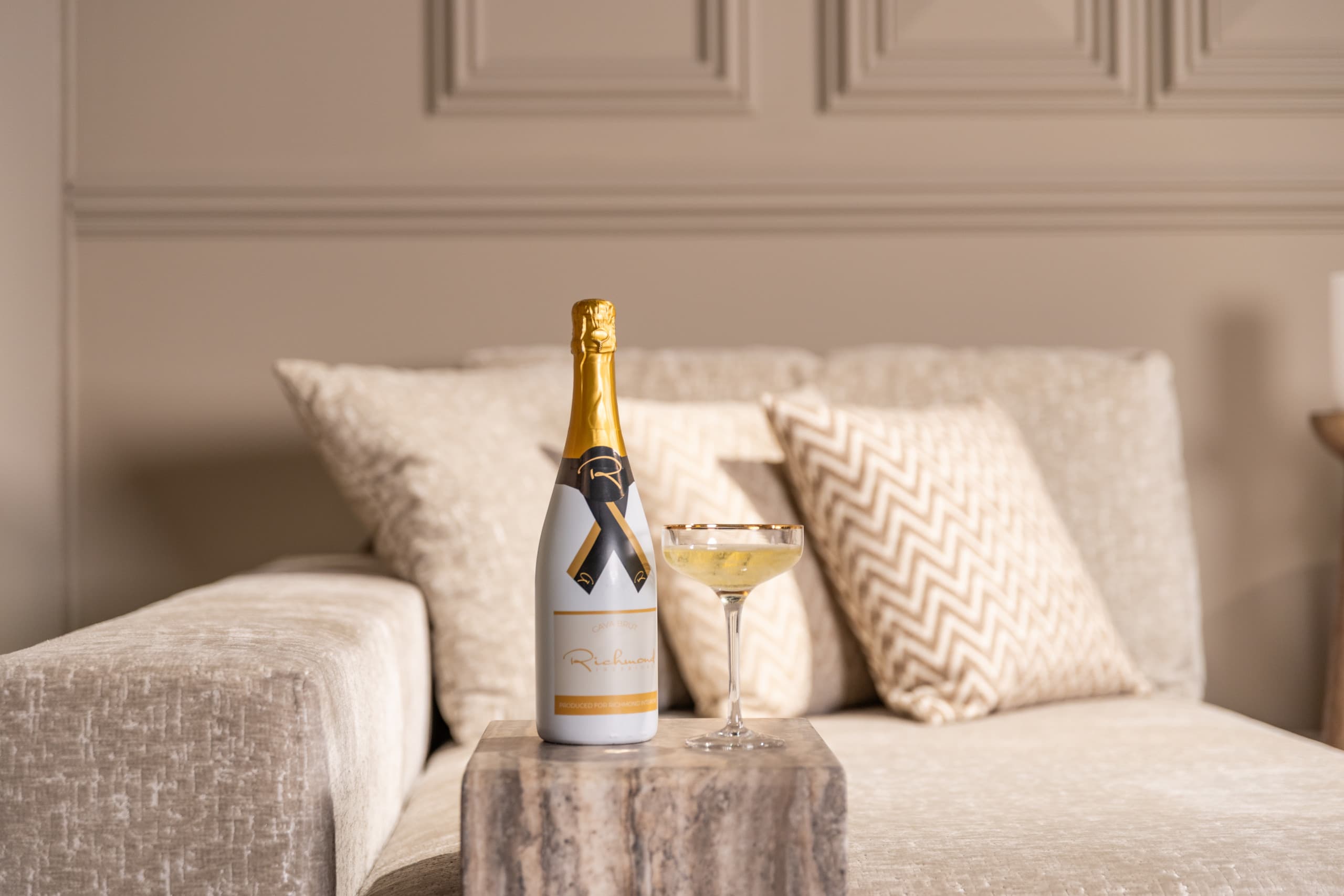 Lichte bank met beige chevronkussens tegen een greige wand, met ervoor een stenen bijzettafel waarop een fles en een glas staan.