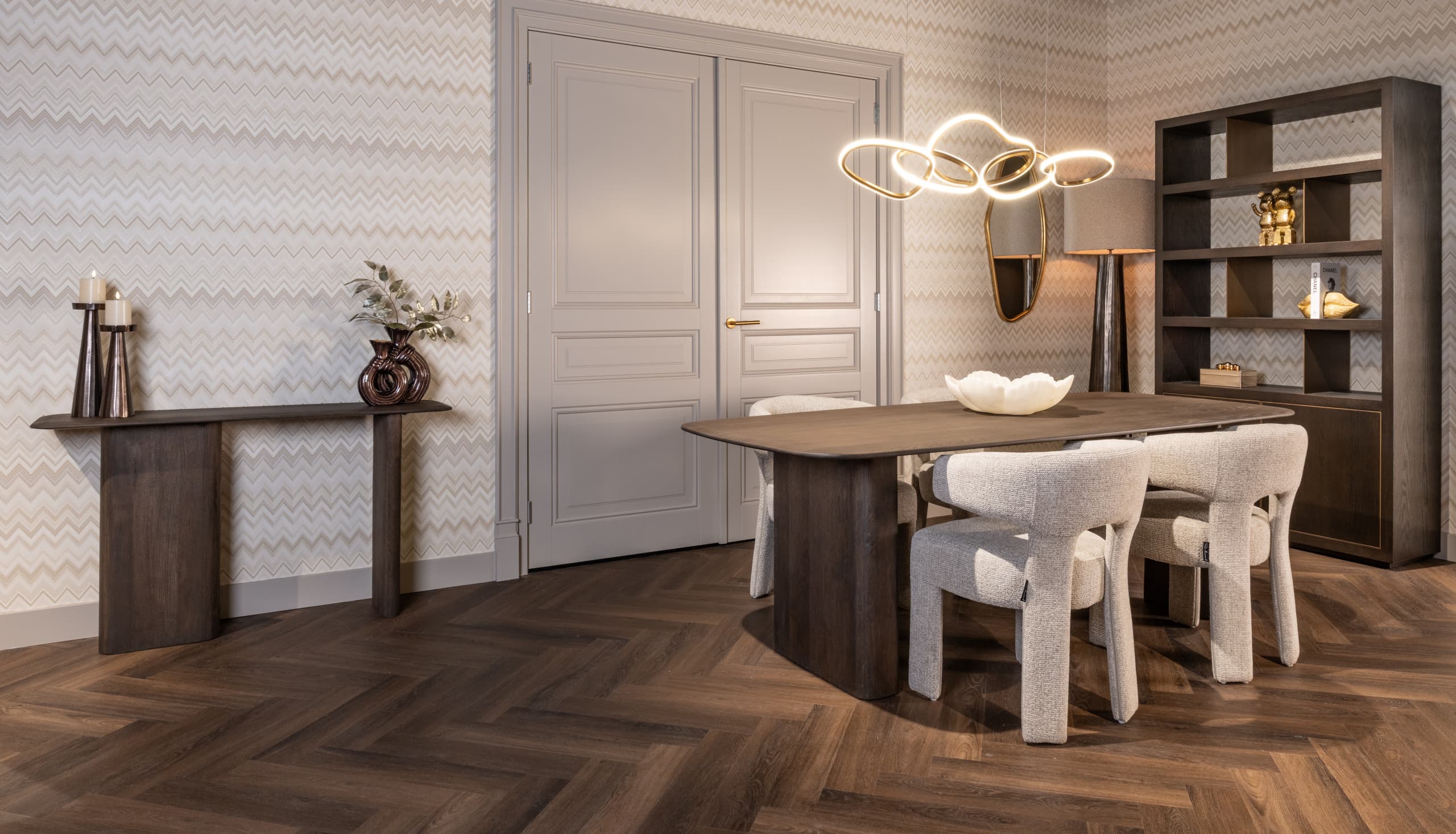Eetkamer met een donkere houten tafel, gouden ringvormige hanglamp, gouden wandspiegel en een donkere vitrinekast met goudkleurige objecten.