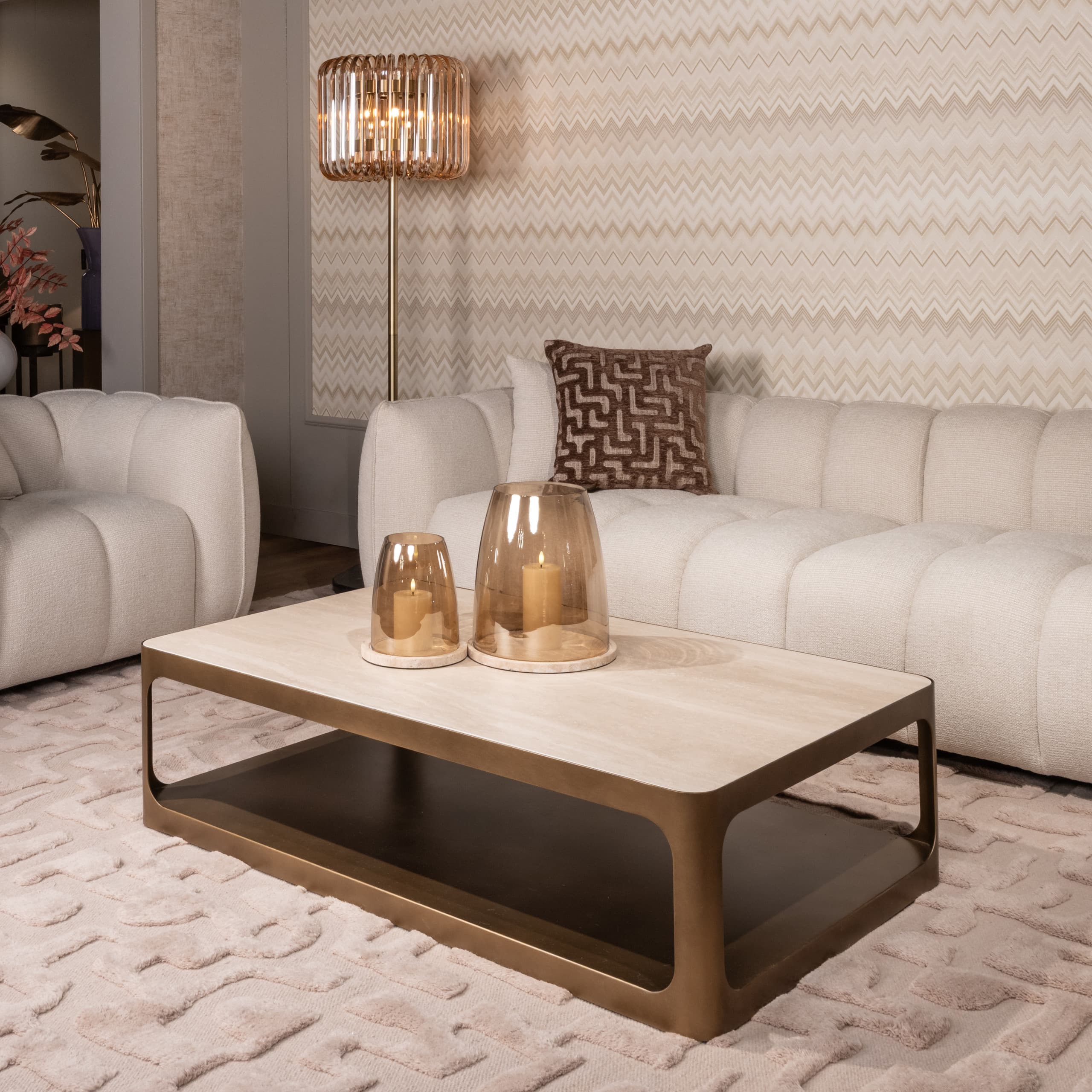 Beige bank met een bruin fluwelen sierkussen en twee brandende kaarsen in amberglazen windlichten op een houten salontafel