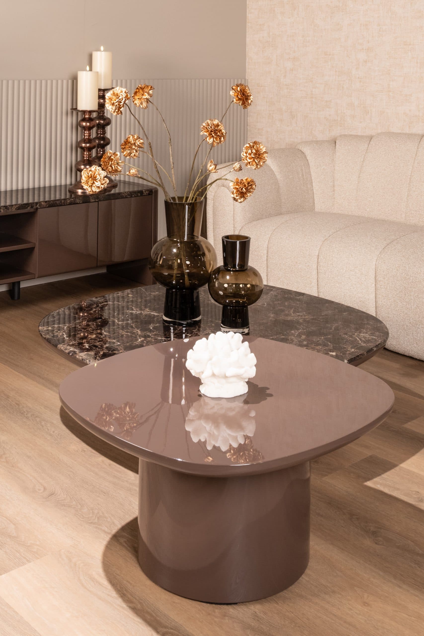 Ovale salontafel met een hoogglans bruin blad en een donker marmeren blad erachter, met twee vazen van gerookt glas, naast een crèmekleurige bank