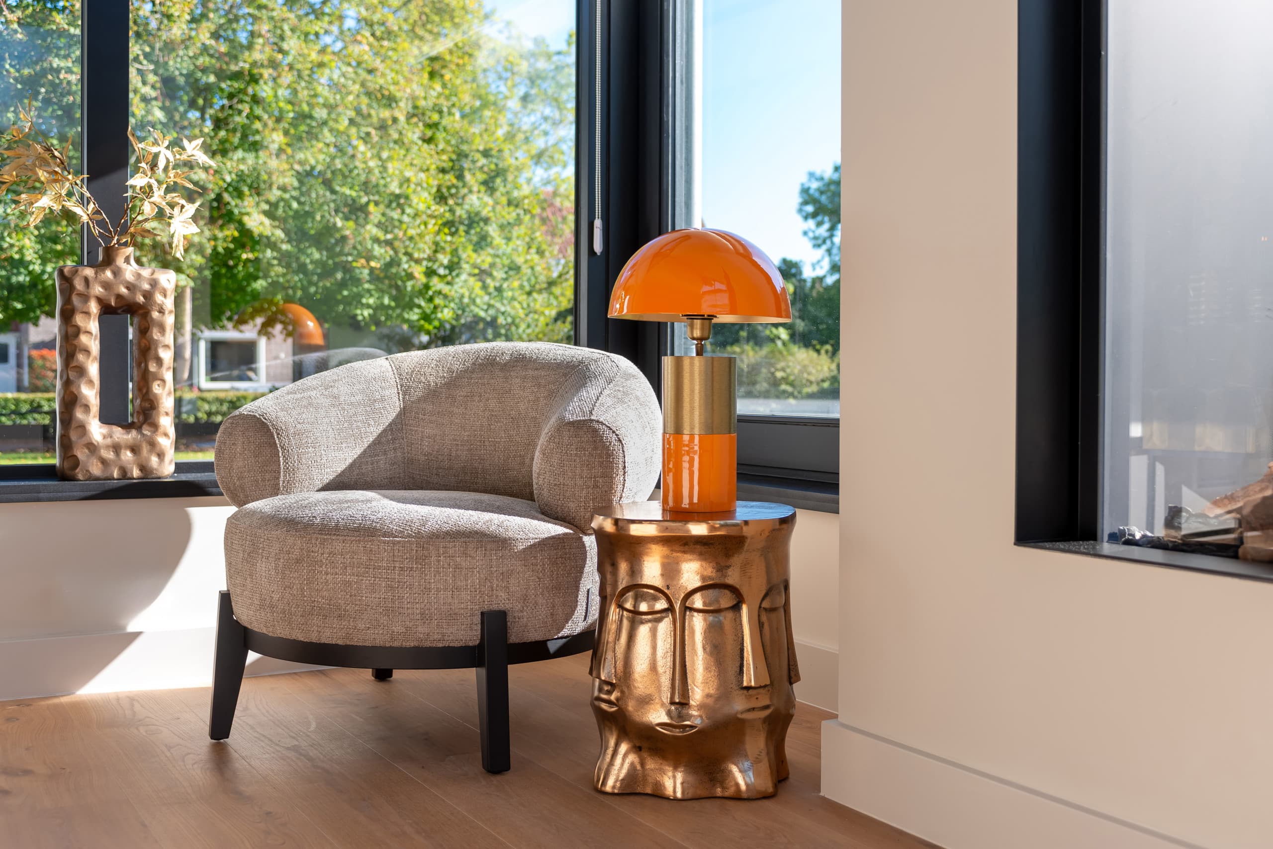 Beige ronde loungestoel op zwarte poten bij een raam, met daarnaast een gouden bijzettafel en een oranje tafellamp