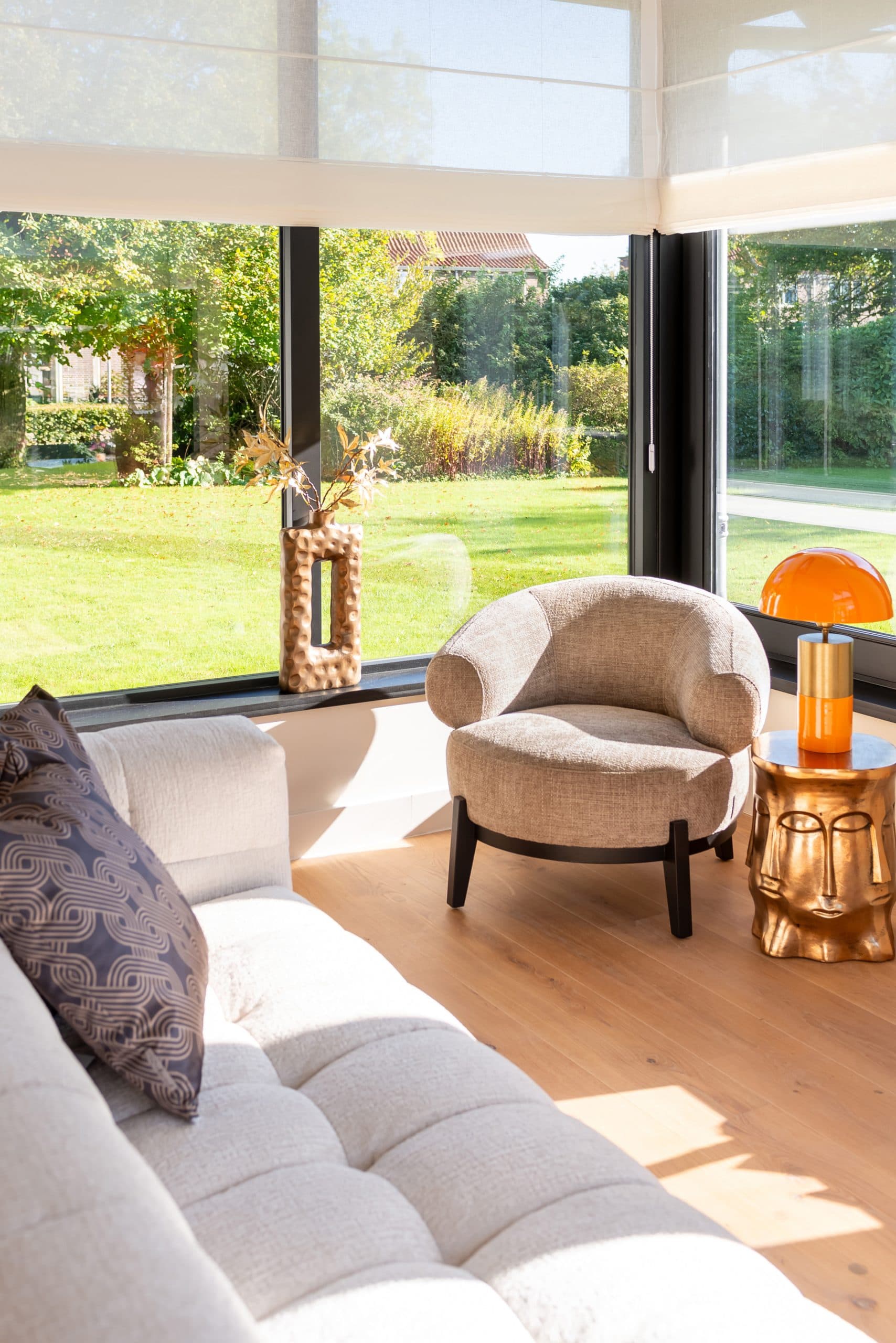 Lichte woonkamer met houten vloer, ronde fauteuil in naturel stof en een lage bank bij grote ramen die uitkijken op een tuin