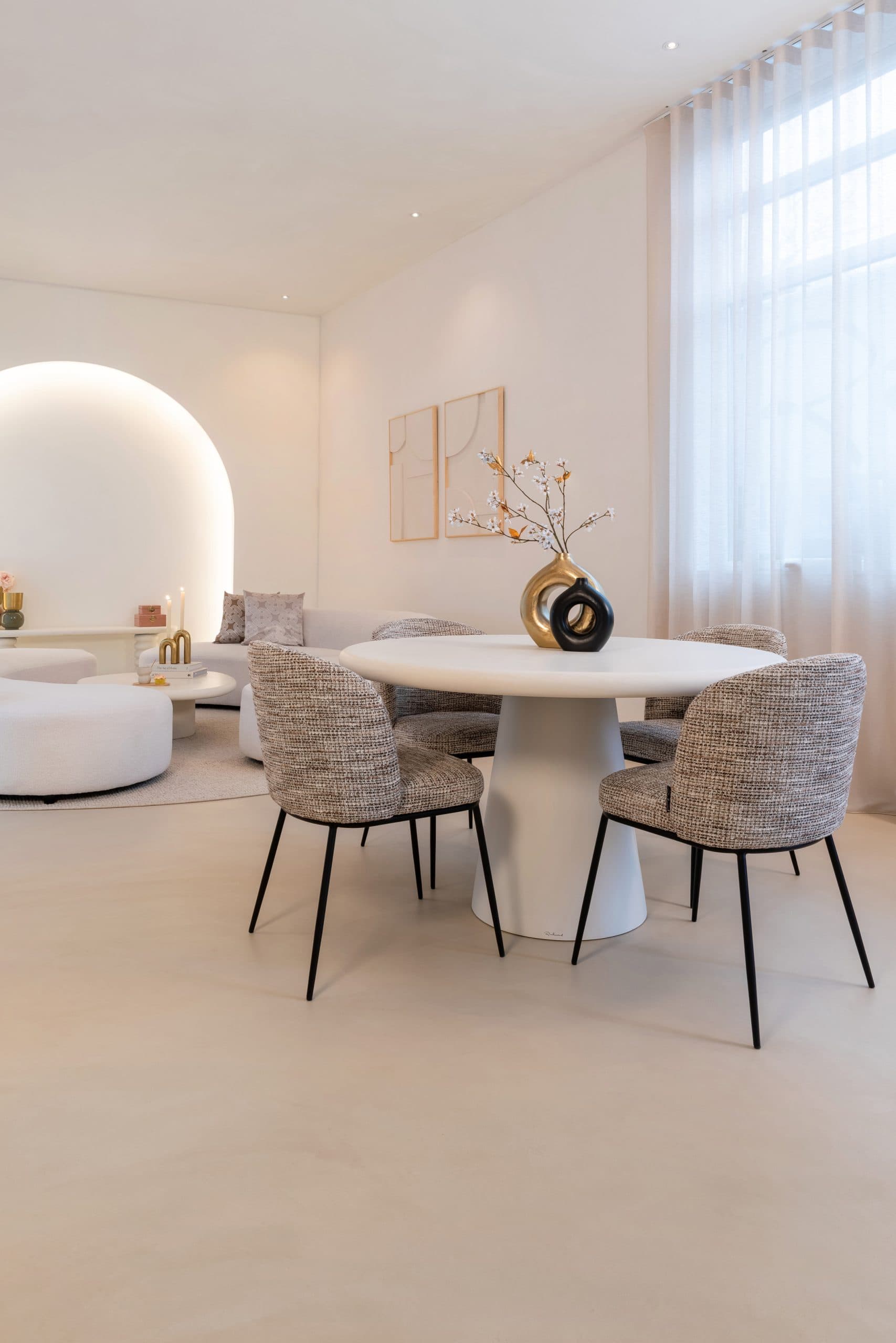 Ronde witte eettafel met vier gestoffeerde stoelen in gemêleerde stof, in een lichte woonkamer met een vaas met bloesemtakken op tafel