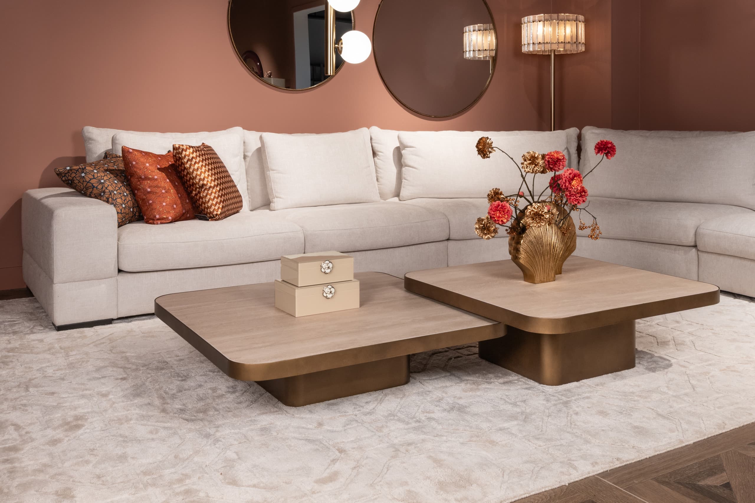 Woonkamer met een lichte hoekbank, twee vierkante salontafels met bronzen voet, een groot vloerkleed en ronde spiegels aan een terracottakleurige wand