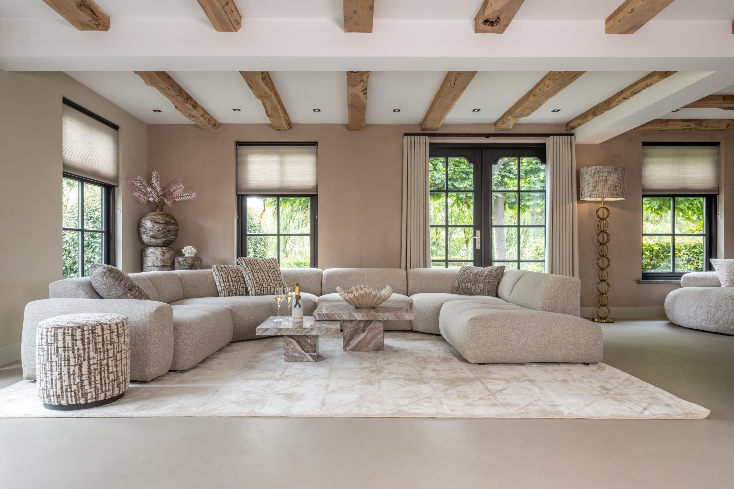 Ruime woonkamer met houten balken in het plafond, een lichte hoekbank, marmeren salontafels, een ronde poef en een vloerlamp bij het raam