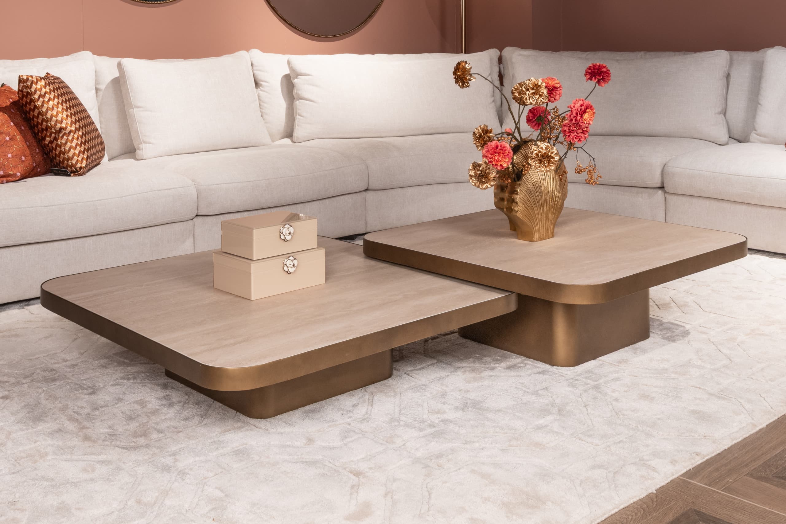 Woonkamer met een crèmekleurige hoekbank, twee vierkante salontafels met houten blad en bronzen voet, een gouden vaas met droogbloemen en twee opbergdoosjes, op een licht vloerkleed