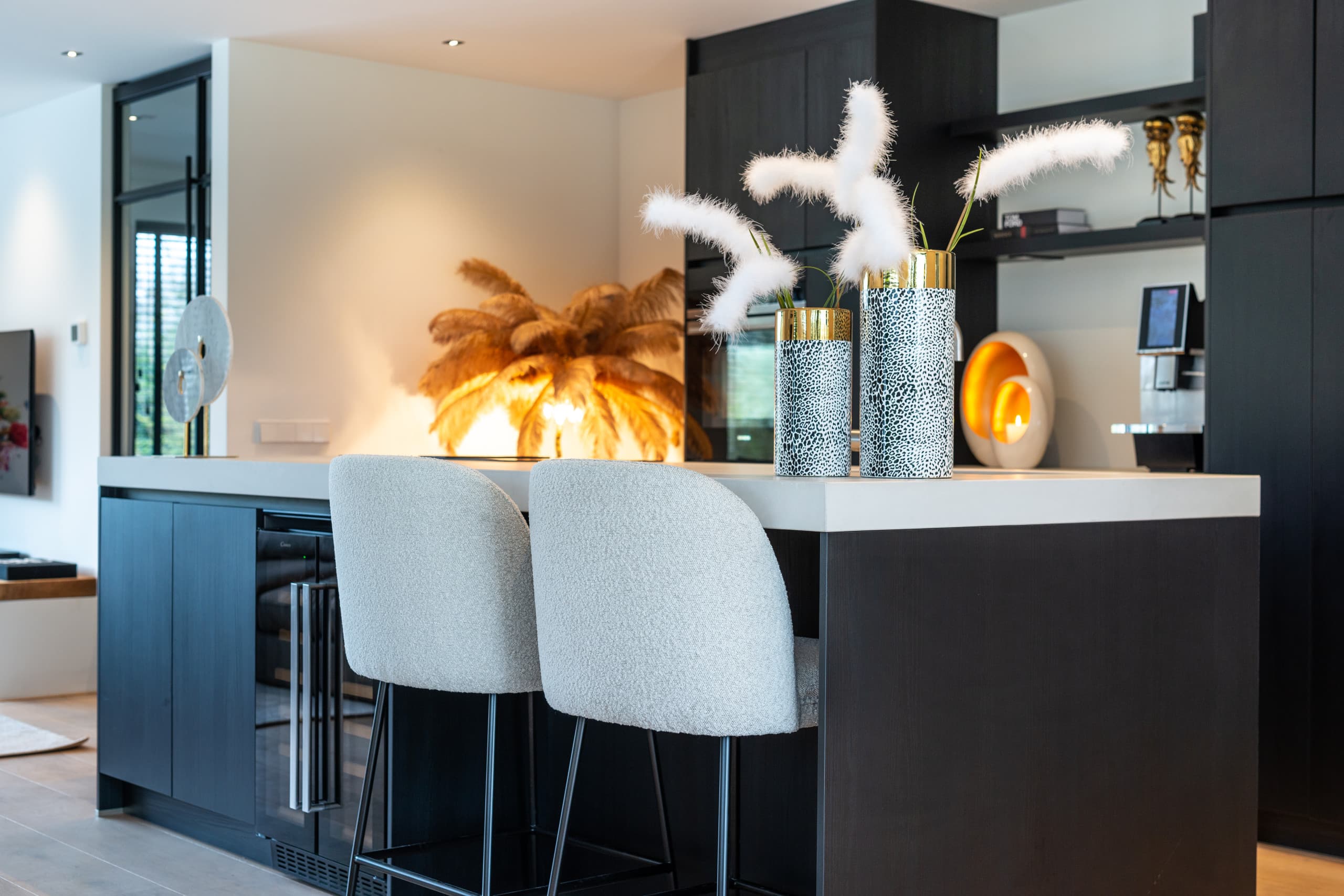 Keukeneiland met twee lichte barkrukken van bouclé, twee gespikkelde vazen met pluimen en een brandende schemerlamp op de achtergrond