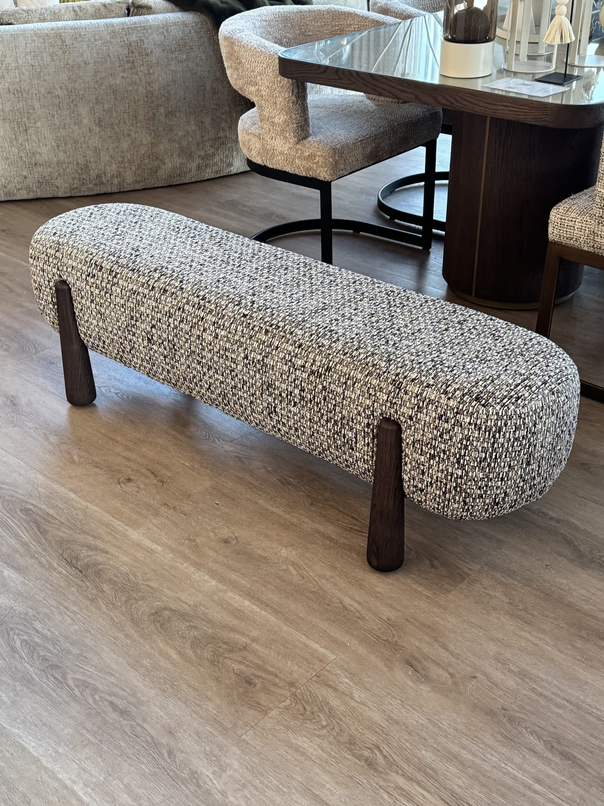 Langwerpig bankje met gemêleerde bouclé zitting op donkere houten poten, op een lichte houten vloer
