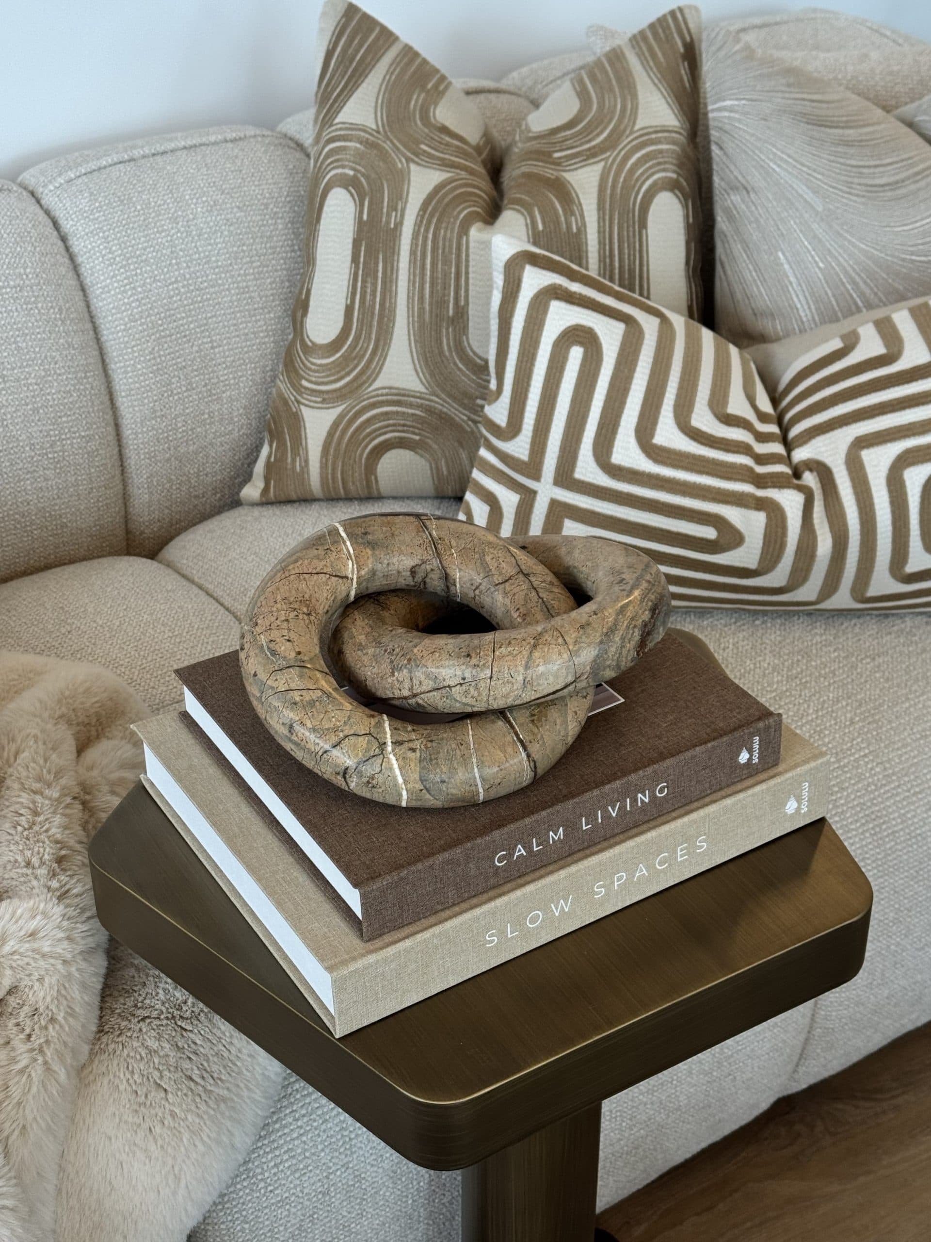 Bruin marmeren knoopsculptuur op een stapel linnen boeken, voor een bank met gedessineerde kussens en een schapenvacht