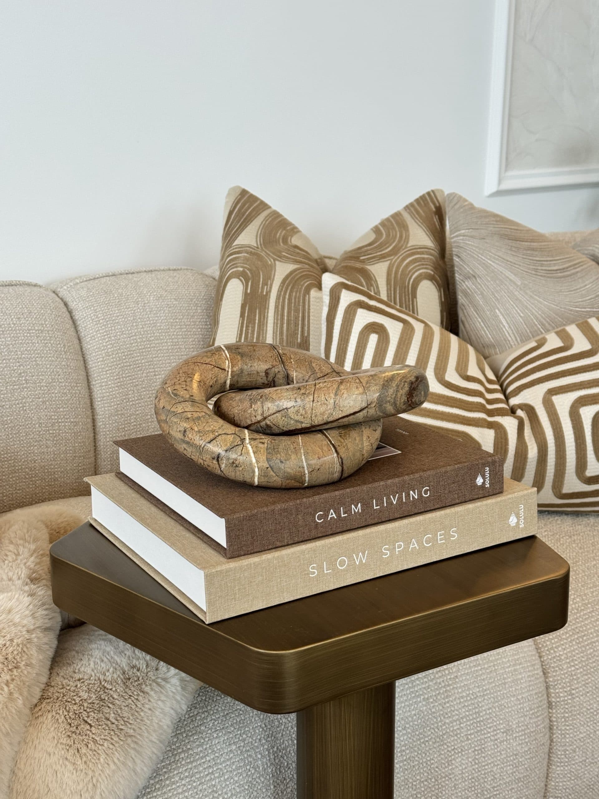 Bruine marmeren knoopsculptuur op een stapel boeken, voor een bank met beige en bruin gedessineerde sierkussens.