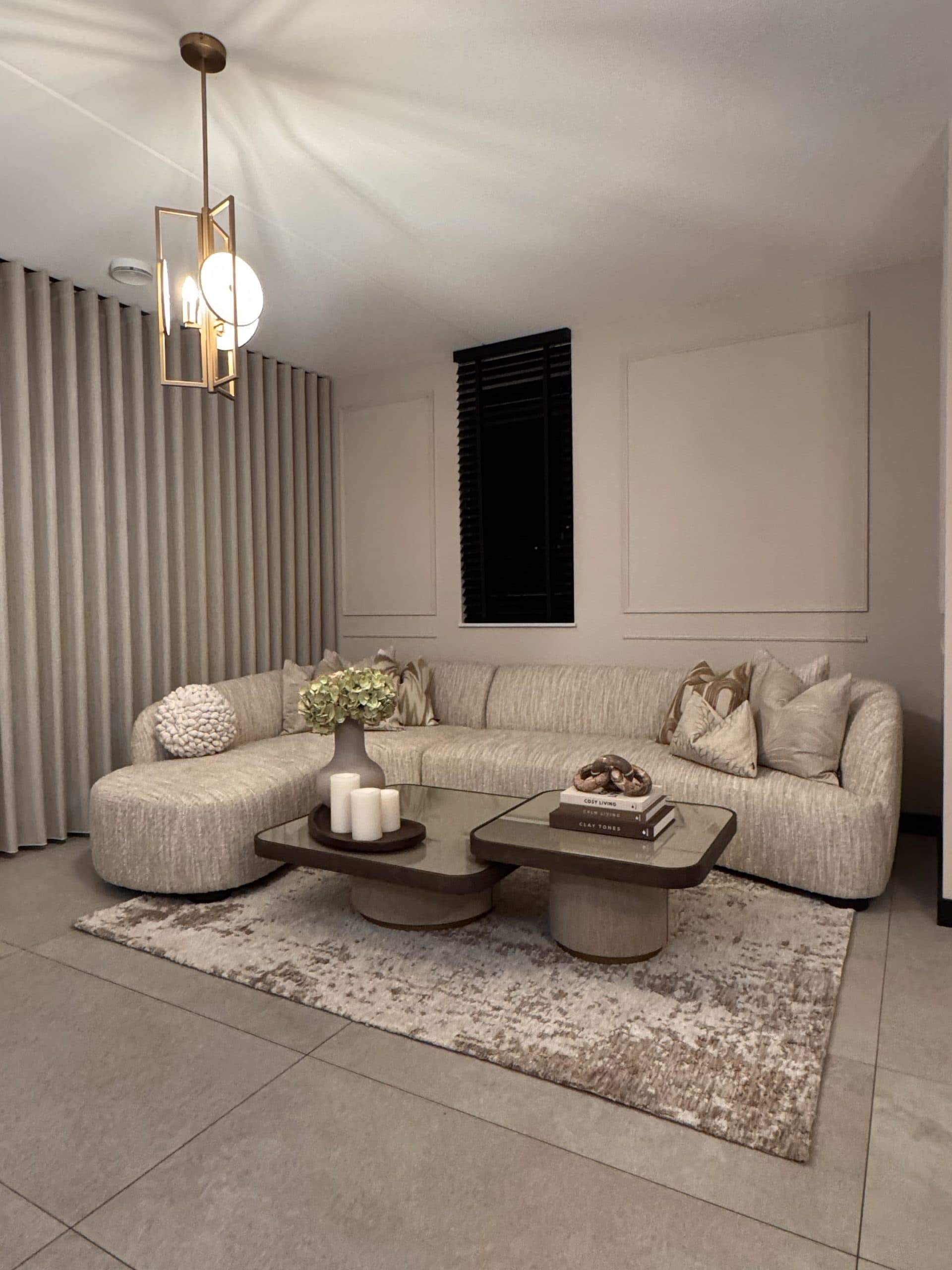 Woonkamer met beige hoekbank vol sierkussens, wandpanelen, lange gordijnen en een salontafel met kaarsen, boeken en een vaas met hortensia's.